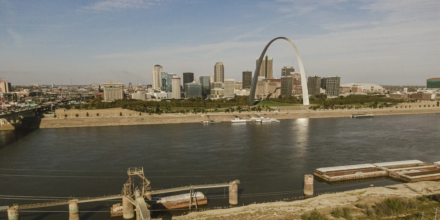 City skyline with a prominent arch next to a river, with boats docked along the waterfront under a partly cloudy sky.