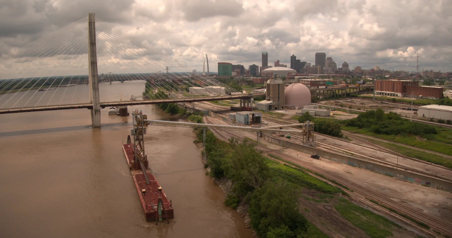 A cable-stayed bridge crosses a river near a city skyline under cloudy skies, with industrial areas nearby.