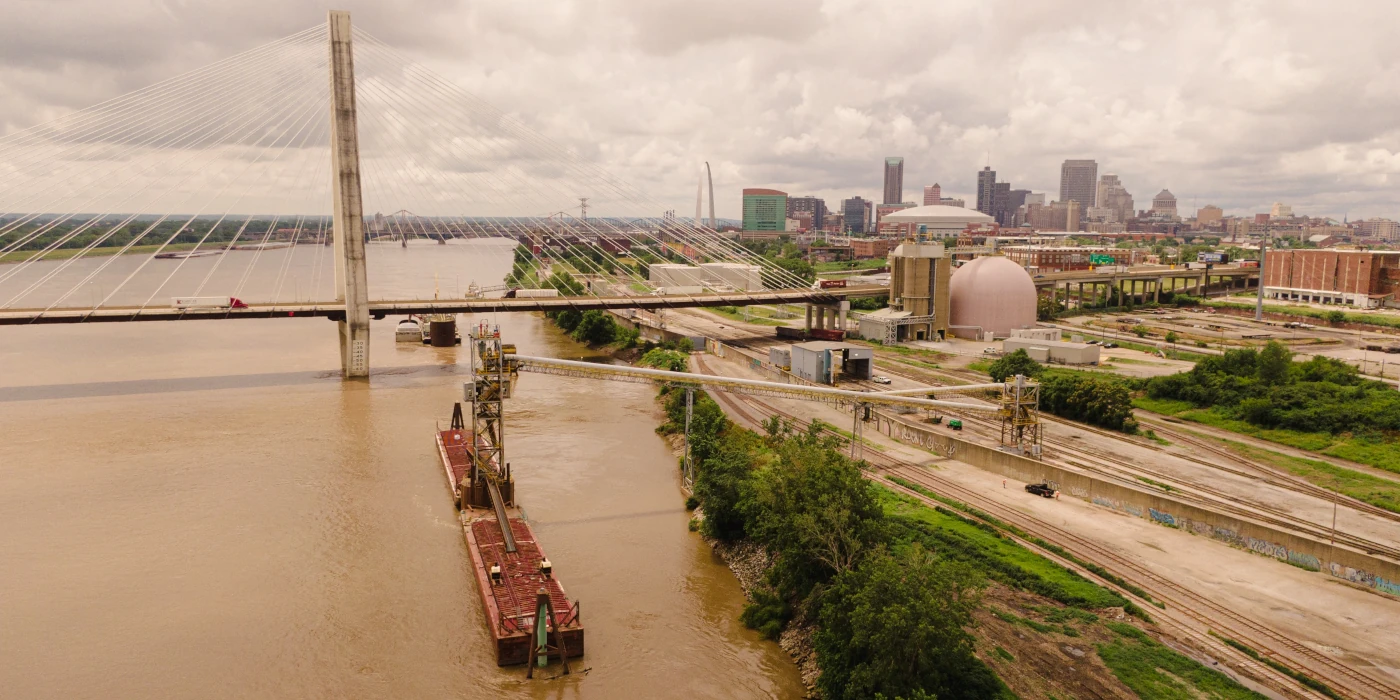 A barge travels on a river beneath a cable-stayed bridge near industrial buildings and a city skyline.