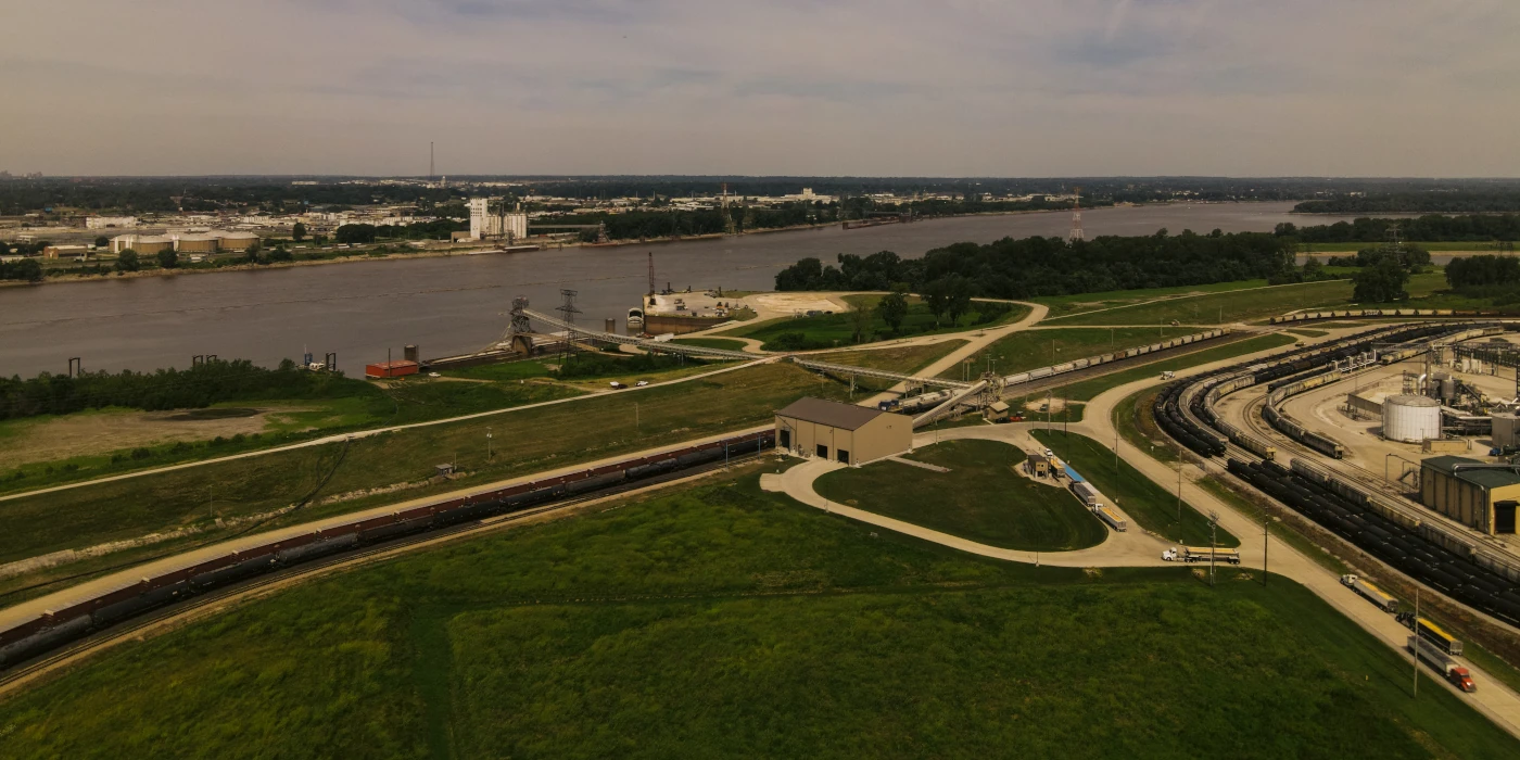 Aerial view of a riverside industrial area with train tracks, storage tanks, and grassy fields.