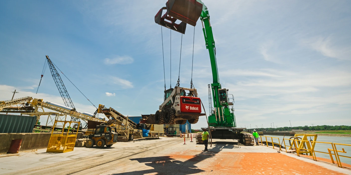 A crane lifts a Bobcat loader at an industrial port with workers standing nearby under a clear sky.
