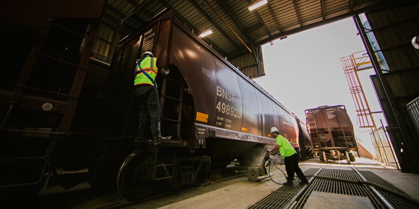Two workers in safety vests inspect and service train cars inside a large industrial rail facility.